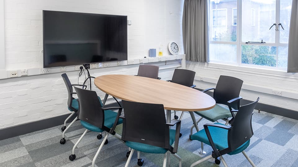 Wavy Top WAV077 - Teaching room with oval table, six chairs, wall-mounted screen, large window, and clock on shelf. Bright, modern setting.