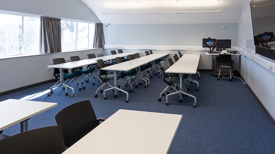 Medium-sized lecture space in Wavy Top, with characteristic curved ceiling, white desks in rows facing forward, white walls and blue carpet.
