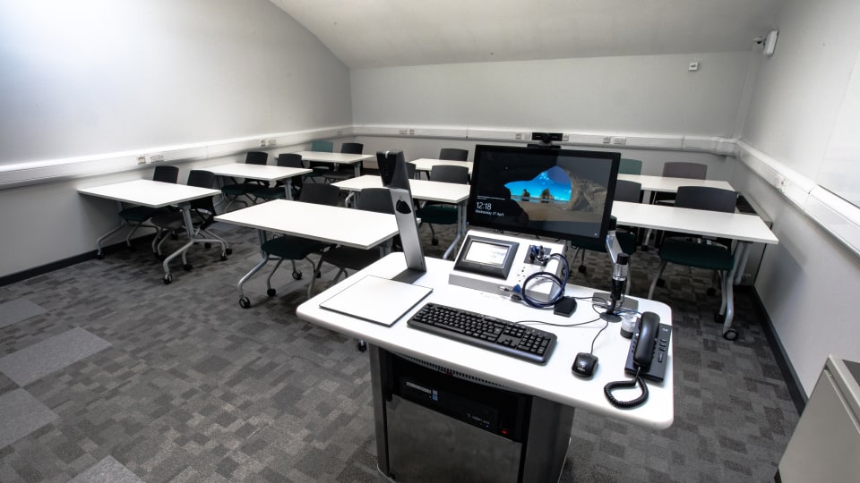 Wavy Top WAV116 - Teaching room with a central lectern featuring a computer, keyboard, microphone, and phone. Rows of tables and chairs fill the space.