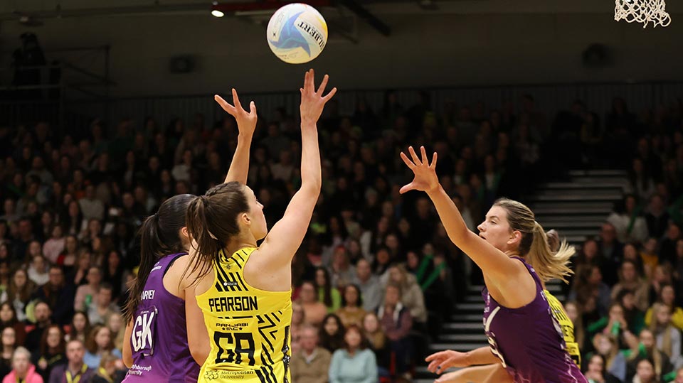 A netball match in progress with players reaching up to catch the ball that is in mid air
