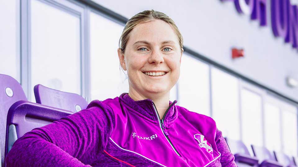 Katie Sorenson wearing a Lightning quarter zip, sitting in the stand at the stadium, smiling whilst looking directly at the camera