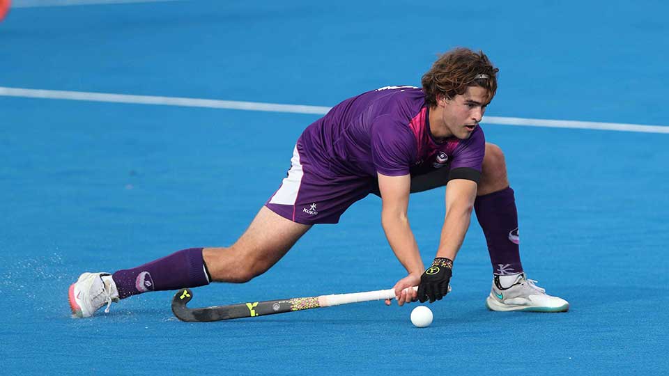 Student, Rory Penrose on a hockey pitch holding a hockey stick, leaning into strike the ball.