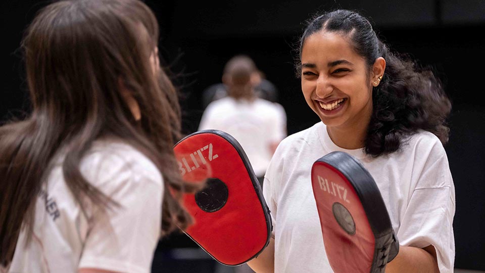 A student wearing boxing pads, holding up their hands, whilst smiling and laughing