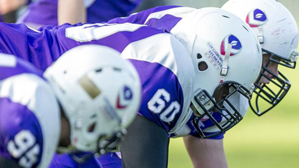 Three American football players in purple and white uniforms and helmets lined up on the field, ready for the next play.