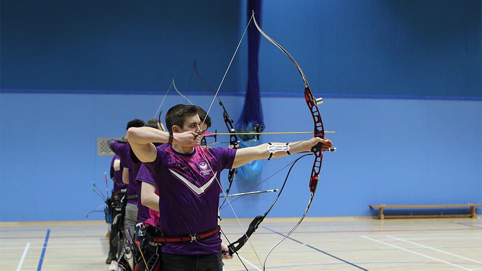 A student in a purple shirt is aiming a recurve bow at a target during an indoor archery session, with other archers visible behind them in a blue-walled gymnasium.