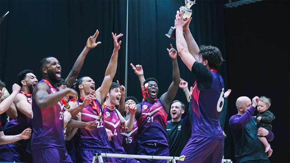 Loughborough basketball team celebrates a championship victory on court, with one player holding a large trophy aloft while surrounded by cheering teammates wearing purple and white jerseys.