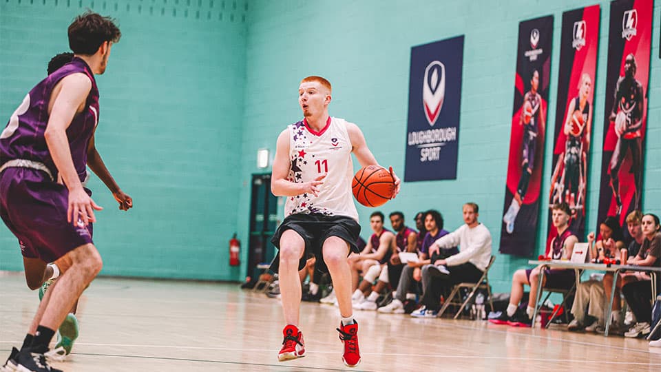 Two male basketball players in action on an indoor court. In the background, spectators sit on the sidelines, and a banner with the 