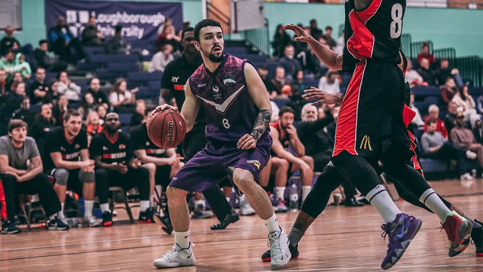 A male basketball player in a purple and black uniform dribbles the ball down the court during a game. He is looking forward, focused on the play, while an opposing player in a black and red uniform guards him. In the background, spectators watch from the stands, and a sign for 