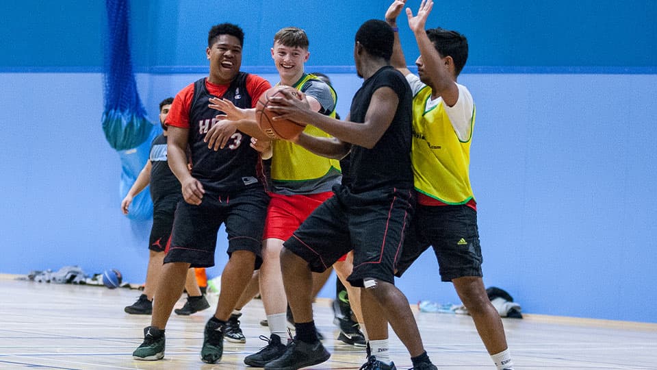 A group of young men in athletic wear playing basketball on an indoor court, with one player dribbling the ball while others are in active play around him.