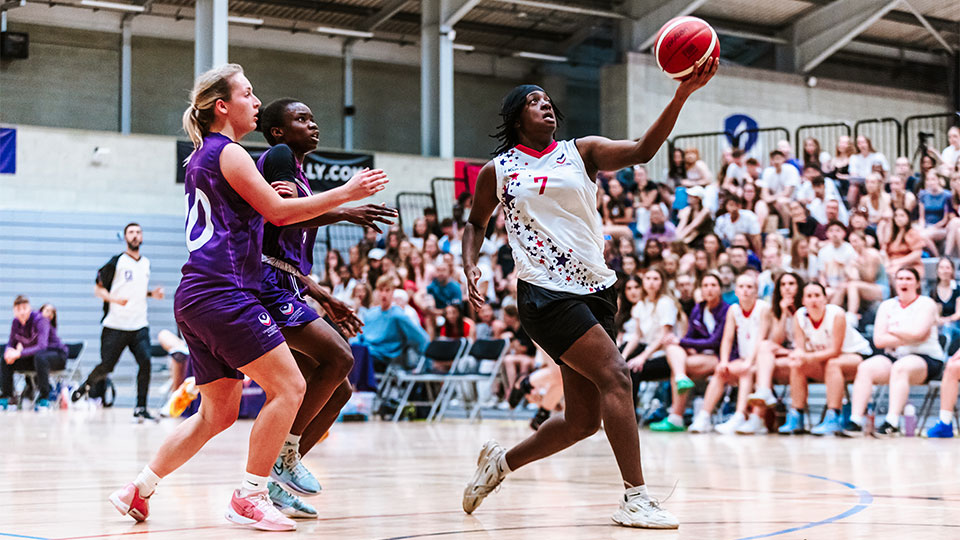 Three female basketball players on an indoor court during a game, with a large crowd visible in the background.
