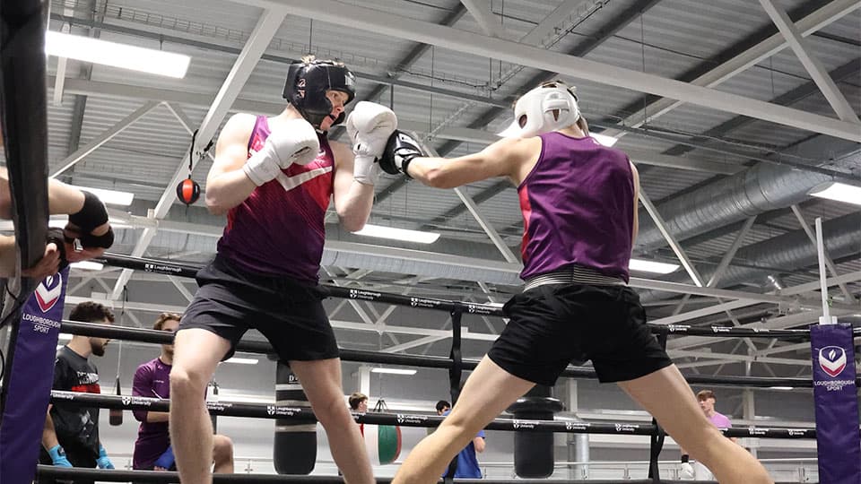 Two male boxers, both wearing black shorts and purple and pink tank tops, spar in a boxing ring. The boxer on the left wears a black head guard and white gloves, while the boxer on the right wears a white head guard and black gloves. Other people are visible in the background, including someone sitting on the ropes of the boxing ring on the far left.