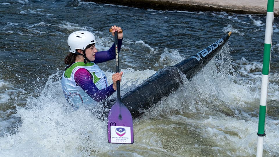 A female kayaker, wearing a white helmet and a purple and orange long-sleeved jacket, paddles through a rapid river.