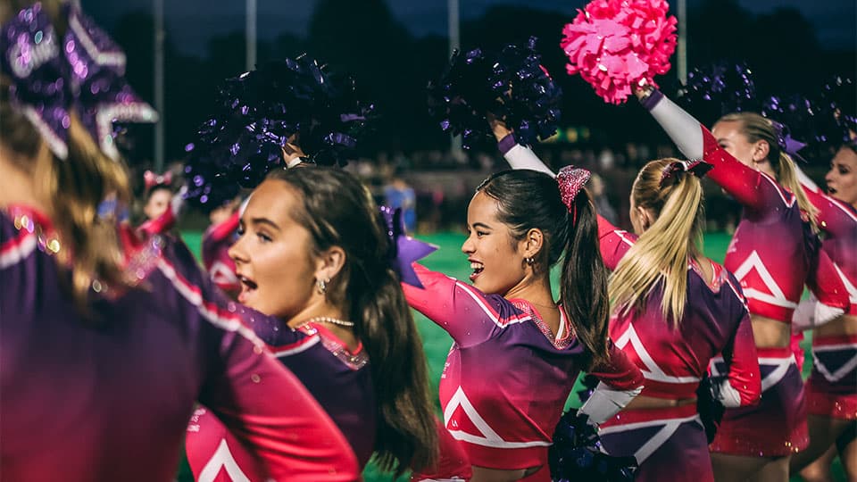A group of cheerleaders, dressed in purple and pink uniforms, perform on a sports field at night. Several cheerleaders are visible from the torso up, some holding purple and pink pom-poms high in the air.