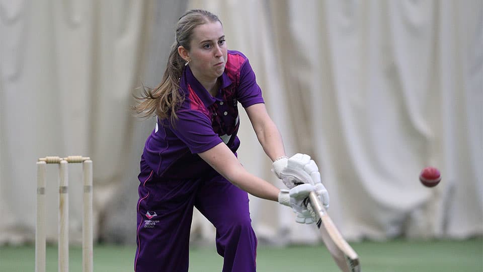 A female cricketer, dressed in a purple uniform, is shown in mid-swing during a practice session.