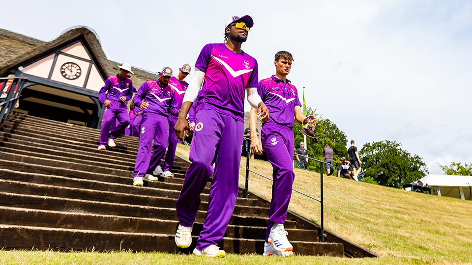 Men's cricket team wearing pink and purple kit walking down concrete steps