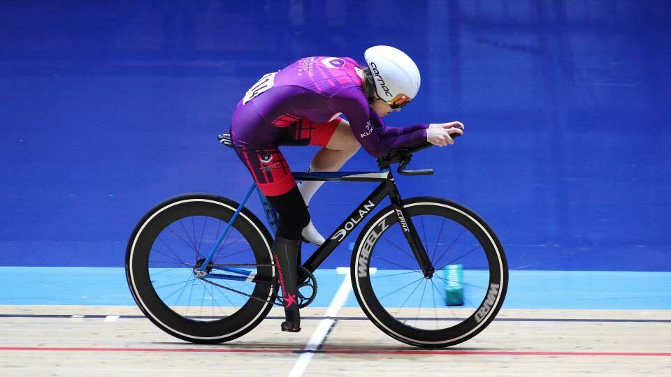 A cyclist in a purple racing suit and white helmet rides a black track bike on an indoor velodrome.