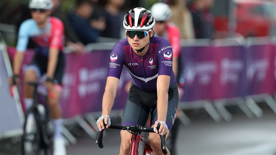 the photo features a cycling event - in the foreground, there is a cyclist wearing university purple cycling gear, some other cyclists appear in the blurred background.