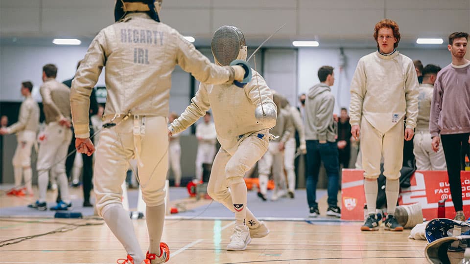 Two people fencing on a wooden floor in a competition hall.
