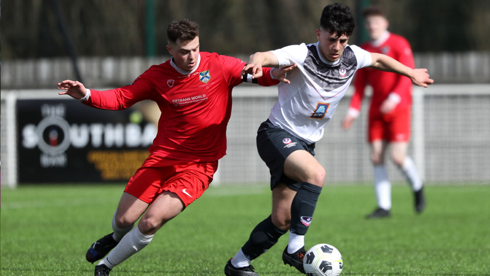 A Loughborough Men's Football player wearing white AU kit, tussling for the ball against an opponent player in all-red kit