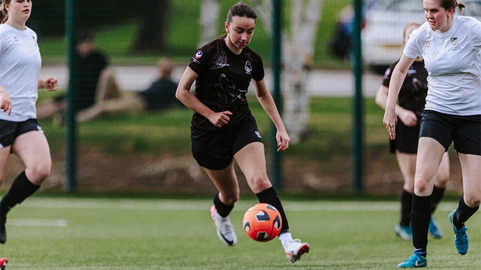 Action shot of a women's soccer match. The player in the centre is running with the football in front of her. Other players visible in the background.