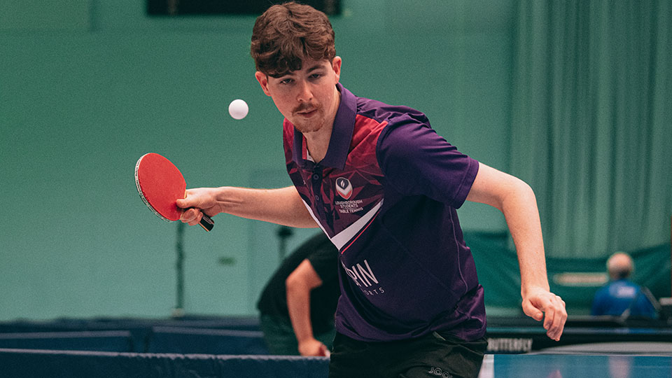 A table tennis player wearing Loughborough kit and about to return the ball.