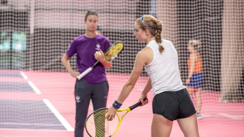 A tennis player on an indoor pink and and purple court preparing to hit the ball during whilst another person is stood watching them.