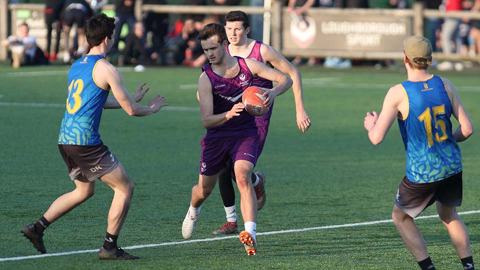 Four touch rugby players on the pitch. Two are wearing purple Loughborough kit and one of them is holding the ball. The other two are wearing a blue kit.