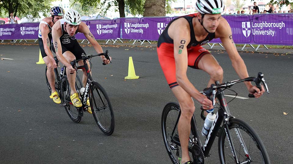 Three triathletes on bikes during a road race. Loughborough University banners are in the background