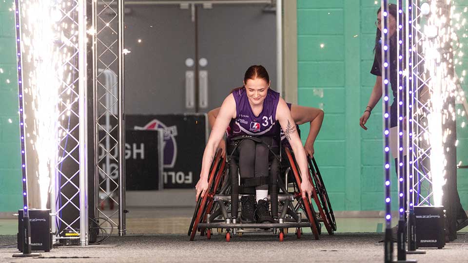 A lightning wheelchair basketball player coming through an archway onto the court. There are sparklers either side of the archway
