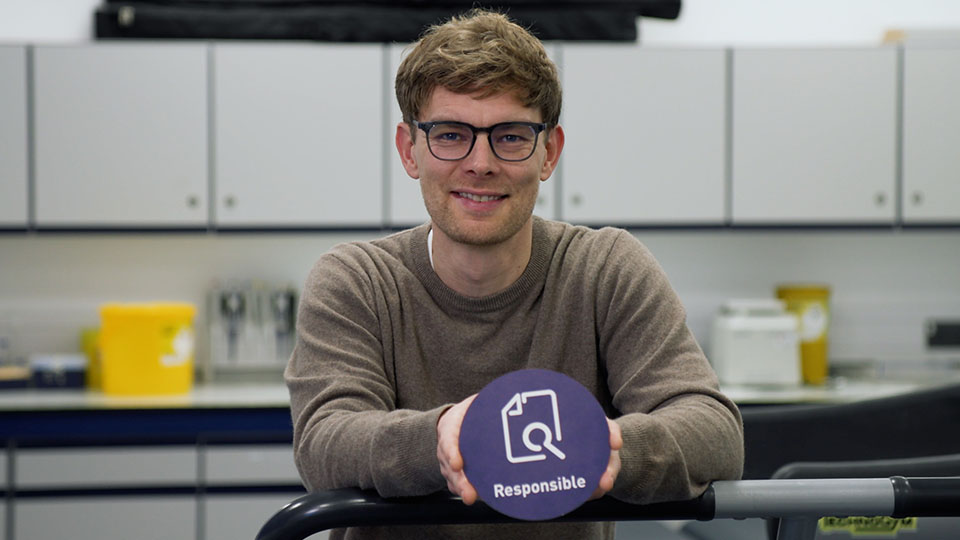Christof in a laboratory wearing glasses looking directly at the camera holding a card circle with an icon and the word 'Responsible' printed on it