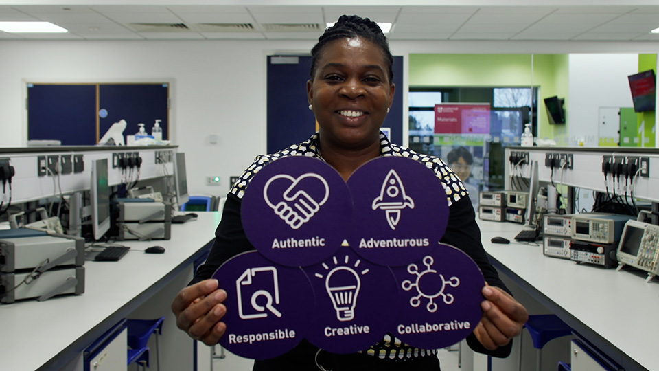 Sheryl standing in a laboratory looking directly at camera holding five card circles icons on them