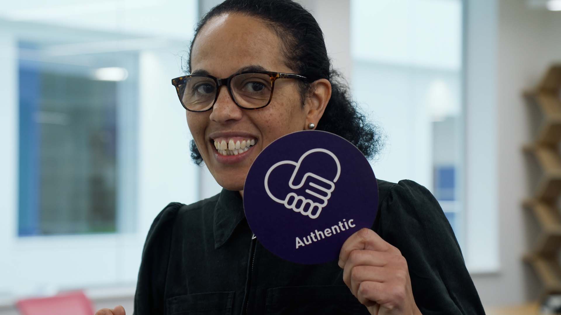 A woman wearing glasses holding a purple card circle with the word Authentic printed on it