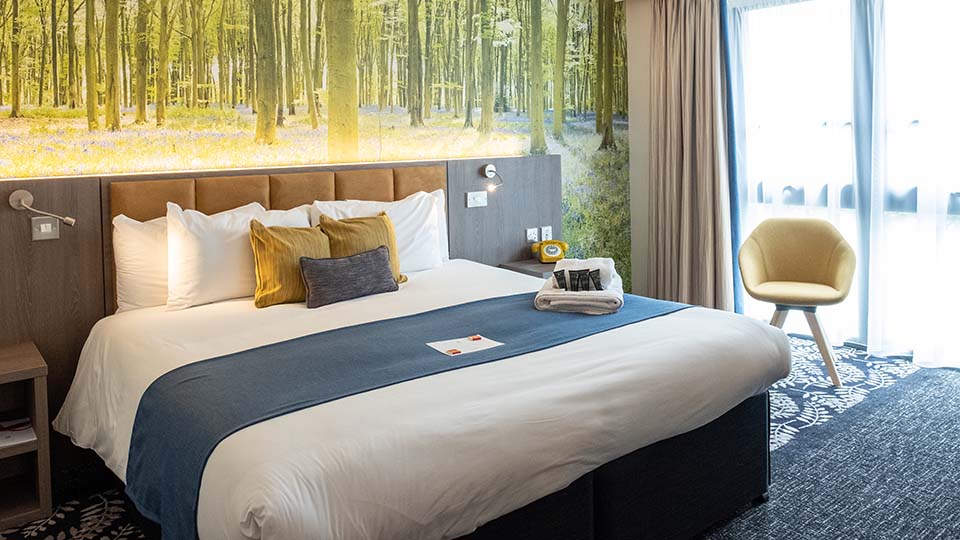 View of a double room: A modern dark carpet, backed with a forest view on the wall behind the bed, which is covered in white linen, a blue bed topper with blue and yellow cushions; yellow chair to the right.