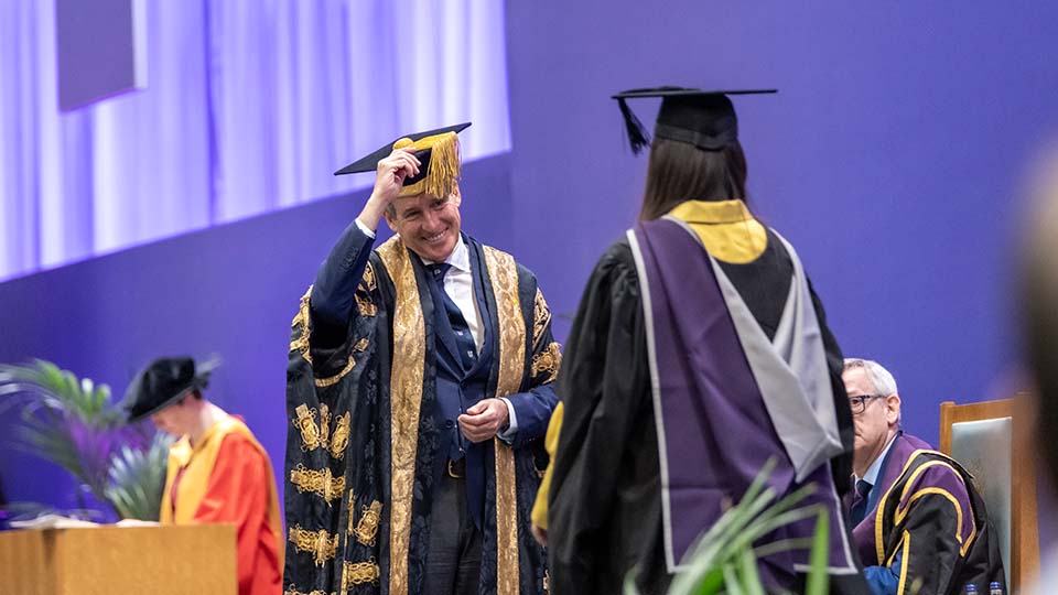 Graduand crossing the stage being greeted by a smiling Chancellor, who is doffing is cap at her.