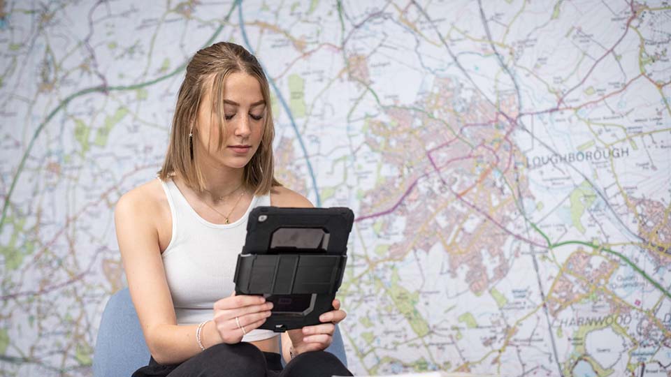 Female student in a white top sat in front of a wall map, intently looking at an iPad.