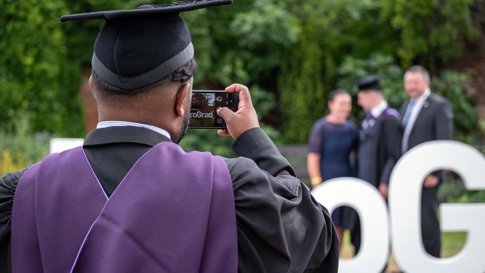 Graduate in gown and mortar taking a photo of another graduate with parents outdoors.