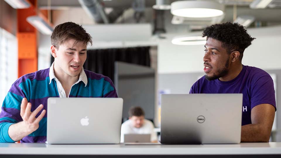Two male students, one talking animatedly referencing his laptop, while another wearing a Team Loughborough t-shirt listens intently.