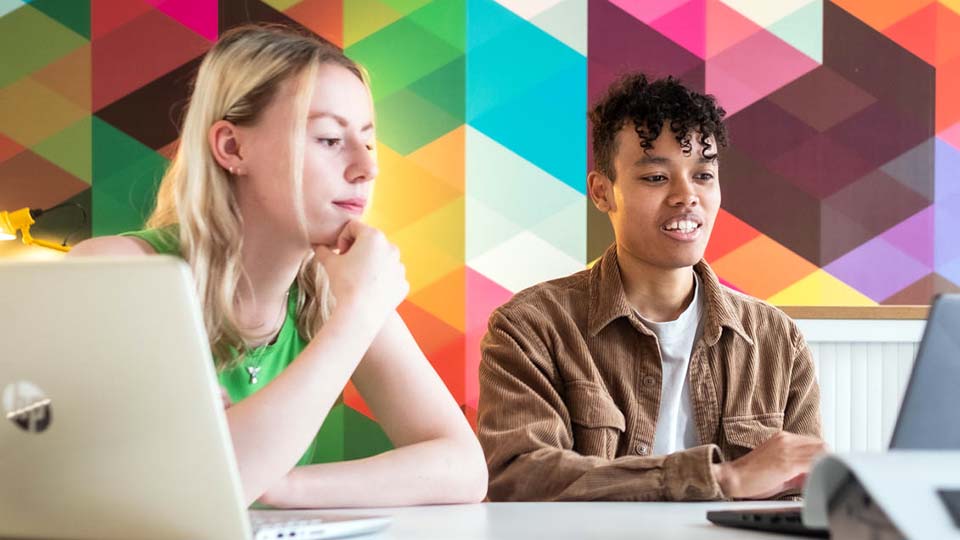 Two students consulting content intently on a laptop, set against a vibrant workspace wallcovering.