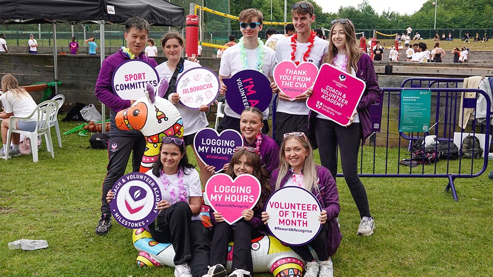 A group of young volunteers at an outdoor event pose together on the grass, smiling and holding colourful signs such as 'Volunteer of the Month', 'Logging My Hours', 'Sport Committees', and 'Thank You from the CVA'. They are part of the Coach & Volunteer Academy at Loughborough University.