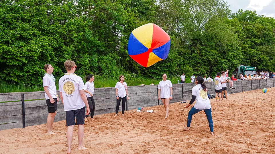 a group of students playing with a large, inflatable ball, barefoot on the sand