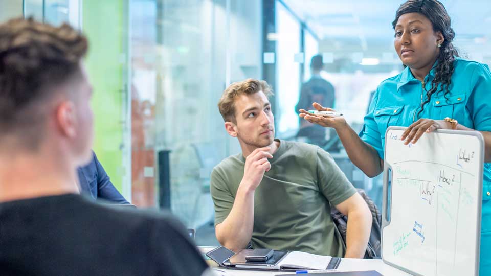 three students, two sitting at a table, one standing holding a whiteboard