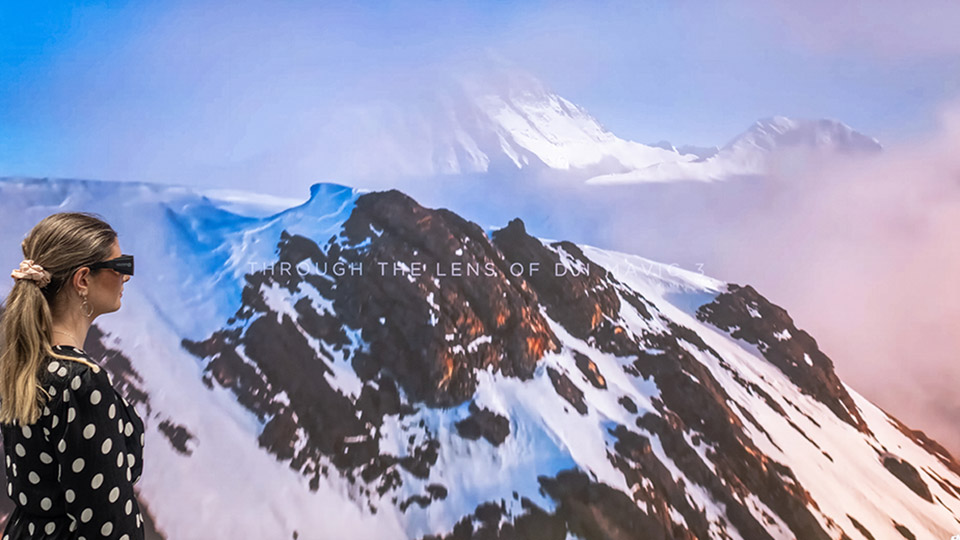A person wearing 3D glasses looking at a large visualisation wall displaying mountain views.