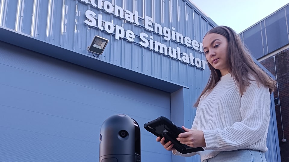 A student holding a tablet next to a laser scanner in front of the National Engineering Slope Simulator facility