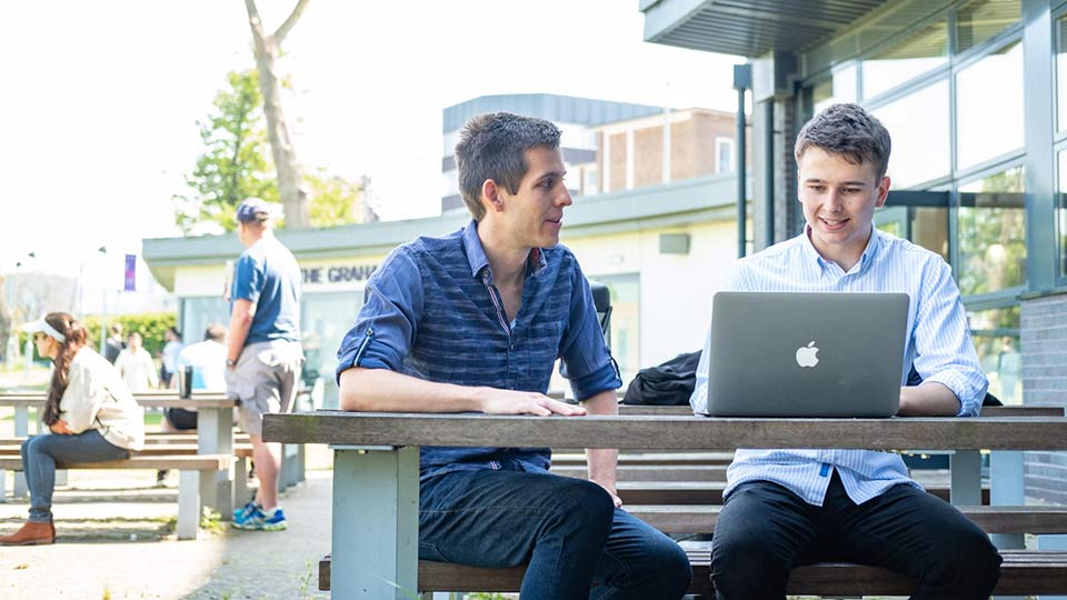 two students sitting at a table outside looking at a laptop computer