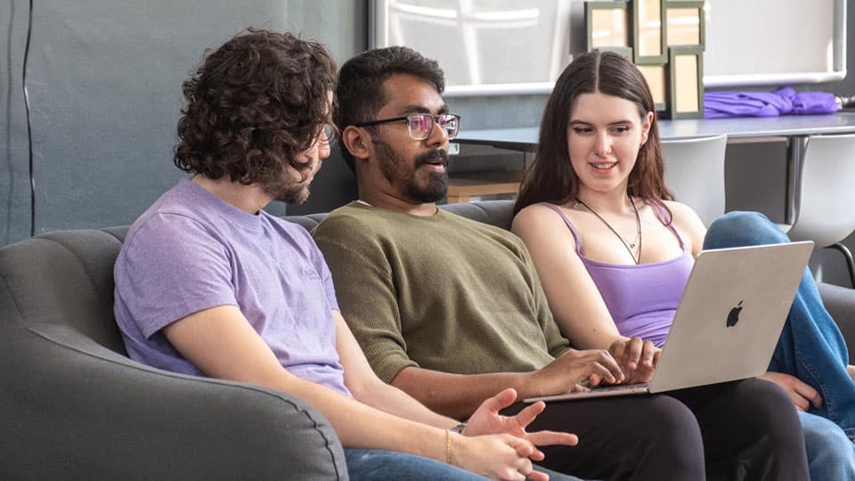 Three students sat on a sofa looking at a laptop