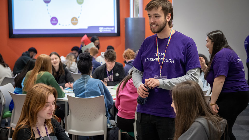 A student ambassador wearing a purple Loughborough t-shirt talking to a group of School students who are sat at tables.