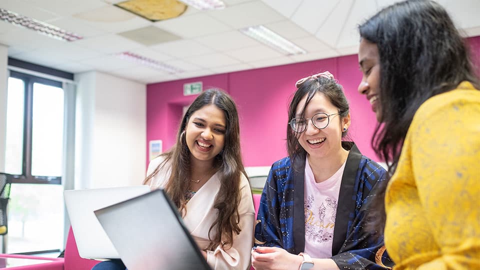 Three female students sitting together on a sofa, looking at each other's laptop screens and smiling