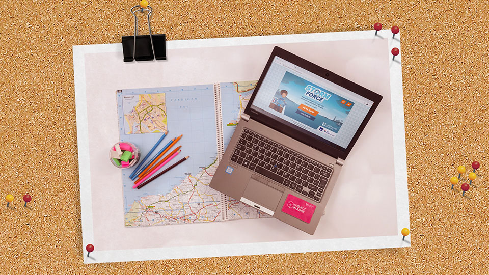 A laptop with the Storm Force game playing on it, on top of a map with some pencils and rubbers to the side.