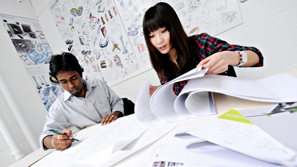 Image of two people looking at their design work. One is sketching, and the other is going through a work book of designs. 