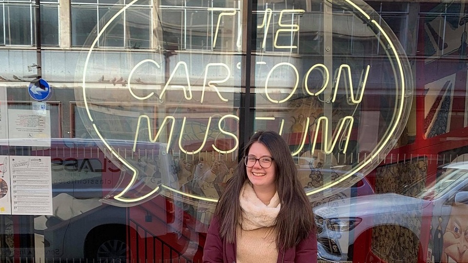 Emma standing in front of an yellow sign of a speech bubble with the words 'the cartoon museum' in it.